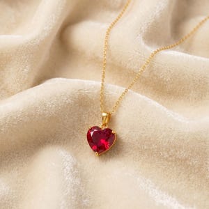May include: A gold-colored necklace with a heart-shaped, ruby-red pendant. The pendant is set in a gold bezel and hangs from a delicate gold chain. The background is a soft, cream-colored fabric.