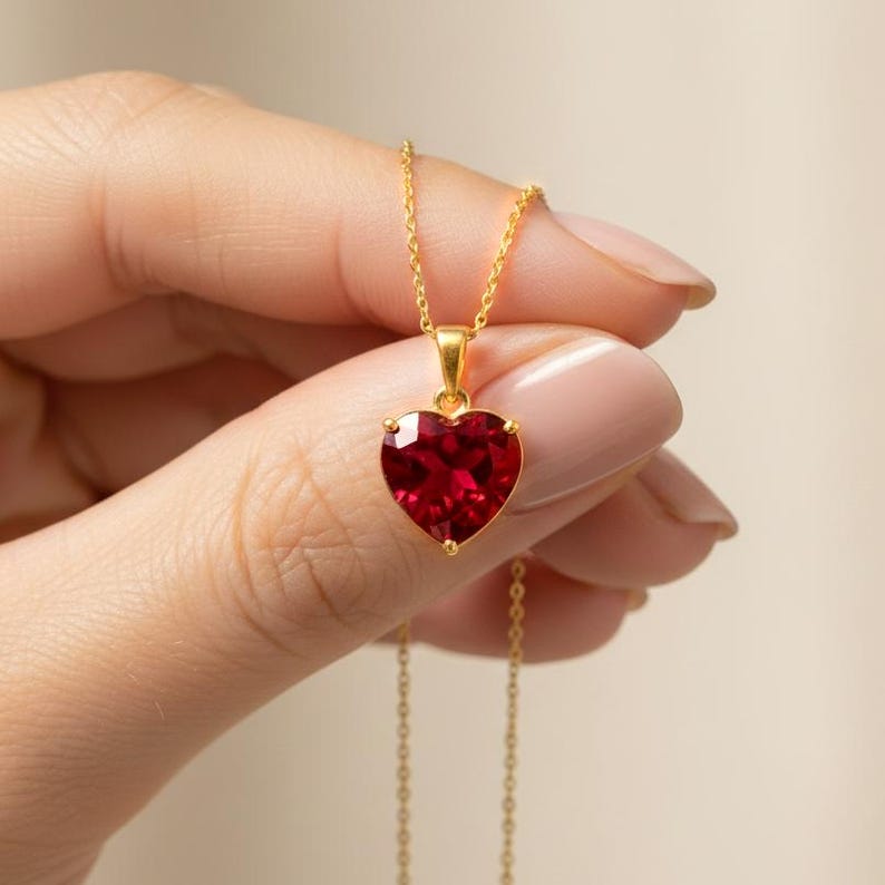 May include: A gold-colored necklace featuring a heart-shaped, deep red gemstone pendant. The pendant is held in a four-prong setting and hangs from a delicate chain. The necklace is a piece of jewelry.