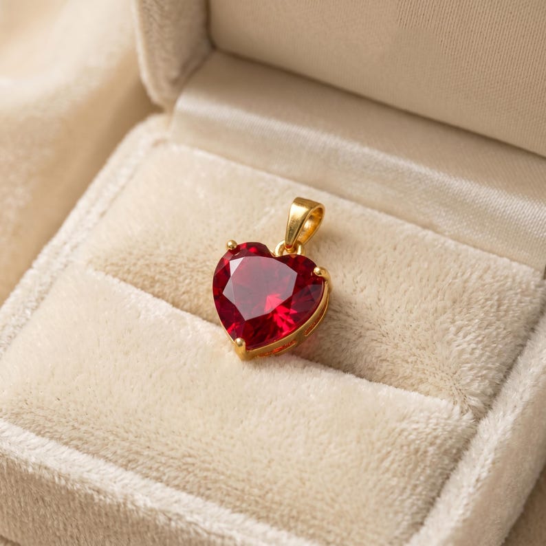 May include: A heart-shaped pendant with a vibrant red gemstone set in a gold-colored metal. The pendant is displayed inside a cream-colored jewelry box, showcasing its elegant design and color contrast.