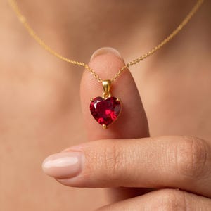 May include: A gold necklace with a heart-shaped, ruby-red pendant. The pendant is held by a person's finger. The necklace chain is delicate and the pendant is set in a gold frame.