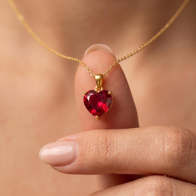 May include: A gold necklace with a heart-shaped, ruby-red pendant. The pendant is held by a person's finger. The necklace chain is delicate and the pendant is set in a gold frame.