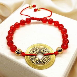May include: Red beaded bracelet with a gold coin charm. The bracelet features a series of red beads and gold accents. The coin charm has Chinese characters and a square hole in the center. The bracelet is on a white surface.