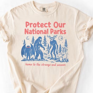 Può includere: T-shirt color avorio con la scritta rosa "Protect Our National Parks". La grafica presenta illustrazioni blu di criptidi e la scritta "home to the strange and unseen".
