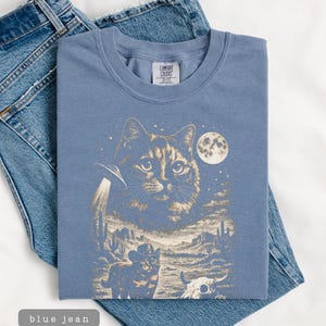 Pu&ograve; includere: Una maglietta blu Comfort Colors con una grafica a tema gatto. Il design include una grande faccia di gatto, un UFO, un gatto cowboy, una luna piena e uno scenario desertico. La maglietta &egrave; appoggiata su un paio di jeans blu.