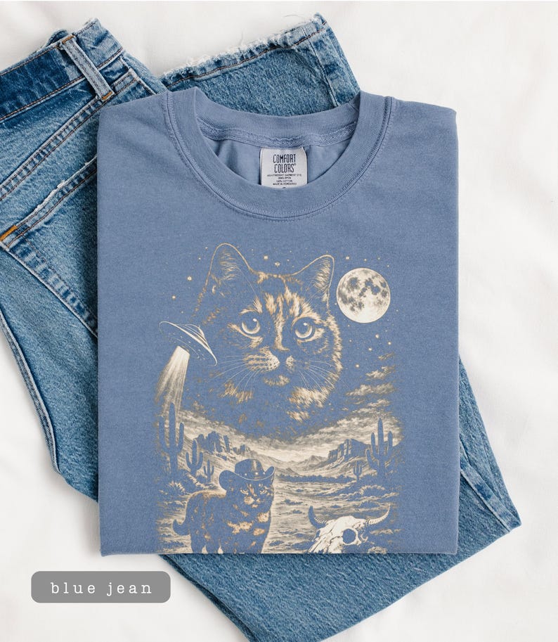 Pu&ograve; includere: Una maglietta blu Comfort Colors con una grafica a tema gatto. Il design include una grande faccia di gatto, un UFO, un gatto cowboy, una luna piena e uno scenario desertico. La maglietta &egrave; appoggiata su un paio di jeans blu.