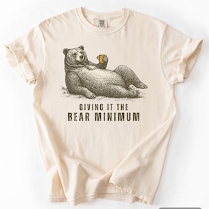 Puede incluir: Camiseta de color marfil con una ilustración de un oso relajado sosteniendo una taza. El texto "GIVING IT THE BEAR MINIMUM" está impreso debajo. La camiseta tiene cuello redondo y mangas cortas.