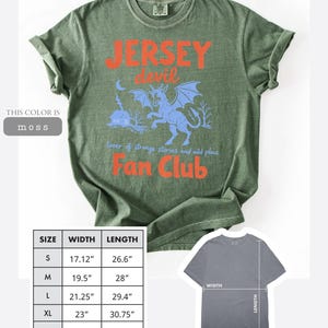 May include: Moss green t-shirt with a graphic featuring the text "Jersey Devil Fan Club" in orange and a blue illustration of the Jersey Devil. The shirt includes size and measurement details in inches.