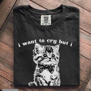 May include: Black t-shirt with a graphic of a kitten wearing a bow tie. The text "i want to cry but i" is printed above the kitten. The shirt is folded on a wooden surface.