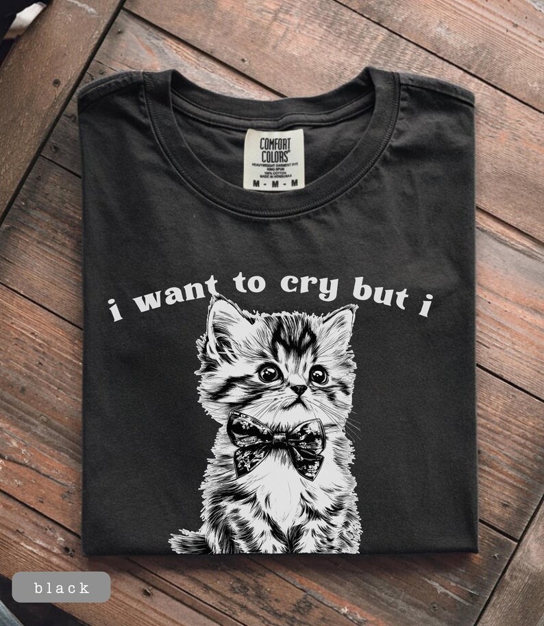 May include: Black t-shirt with a graphic of a kitten wearing a bow tie. The text "i want to cry but i" is printed above the kitten. The shirt is folded on a wooden surface.