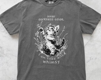 May include: Dark grey t-shirt featuring a black and white illustration of a kitten reaching upwards amidst floral elements. The text "inner darkness aside, I am filled with whimsy" is printed above and below the kitten.