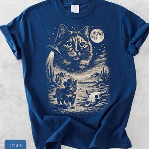 Pu&ograve; includere: T-shirt blu navy con grafica a tema gatto. Il design include un grande volto di gatto, un UFO, un gatto con un cappello da cowboy, una luna piena, uno scenario desertico e un teschio di mucca. Il testo "true navy" &egrave; in un riquadro blu.