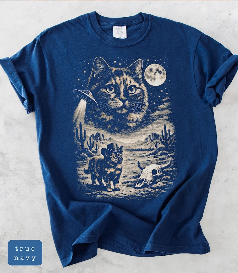 Pu&ograve; includere: T-shirt blu navy con grafica a tema gatto. Il design include un grande volto di gatto, un UFO, un gatto con un cappello da cowboy, una luna piena, uno scenario desertico e un teschio di mucca. Il testo "true navy" &egrave; in un riquadro blu.