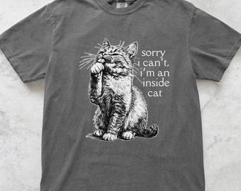May include: A dark gray t-shirt featuring a detailed black and white illustration of a cat. The cat is sitting and appears to be deep in thought. Text on the shirt reads, "sorry, I can't, I'm an inside cat."