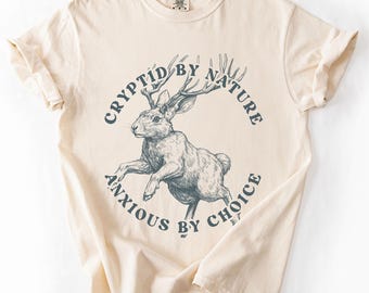 May include: Ivory-colored t-shirt featuring a graphic of a jackalope with antlers, encircled by the text "CRYPTID BY NATURE, ANXIOUS BY CHOICE" in navy blue. The shirt has short sleeves and a crew neck.