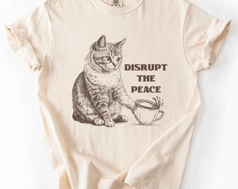 Puede incluir: Camiseta de color marfil con una ilustración en tonos sepia de un gato volcando una taza de café. El texto "DISRUPT THE PEACE" está impreso en marrón. La camiseta tiene cuello redondo y mangas cortas.