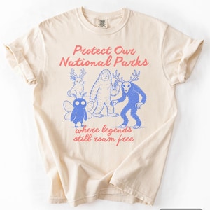 May include: Ivory-colored t-shirt with a graphic design featuring a rabbit, a Bigfoot-like creature, a deer-like figure, and a fairy. The text "Protect Our National Parks" and "where legends still roam free" are printed on the shirt.