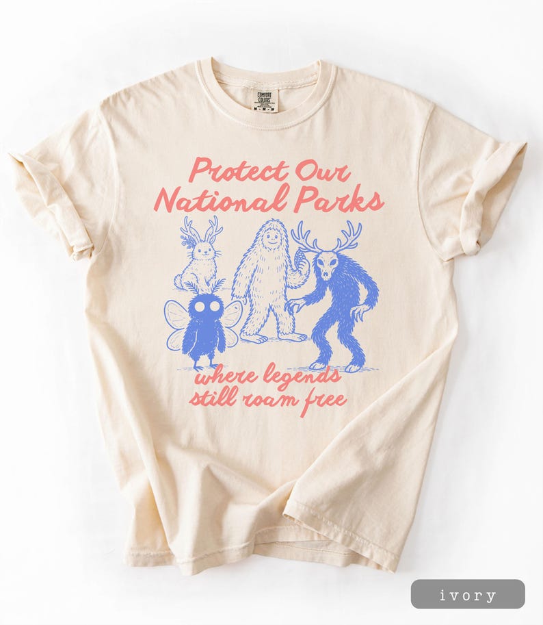 May include: Ivory-colored t-shirt with a graphic design featuring a rabbit, a Bigfoot-like creature, a deer-like figure, and a fairy. The text "Protect Our National Parks" and "where legends still roam free" are printed on the shirt.
