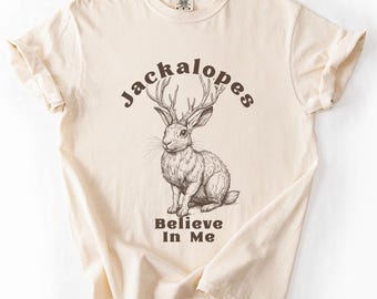May include: Ivory-colored t-shirt featuring a detailed illustration of a jackalope with antlers. The word "Jackalopes" arches above the jackalope, and "Believe In Me" is printed below. The shirt is made of soft material.