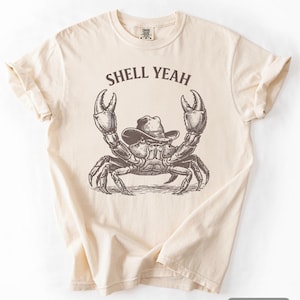 Könnte beinhalten: Elfenbeinfarbenes T-Shirt mit einem Krebs, der einen Cowboyhut trägt, und dem Text "SHELL YEAH". Die Krabbenillustration ist detailliert und im Vintage-Stil gehalten. Das T-Shirt hat einen klassischen Rundhalsausschnitt und kurze Ärmel.