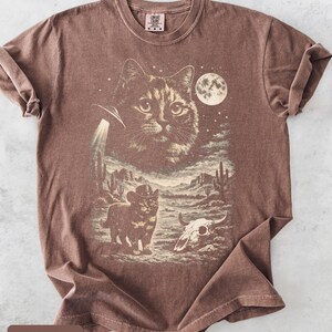 Pu&ograve; includere: T-shirt color espresso con grafica a tema gatto. Il design include un grande volto di gatto, un UFO, un gatto cowboy, un paesaggio desertico e una luna piena. La maglietta ha girocollo e maniche corte.
