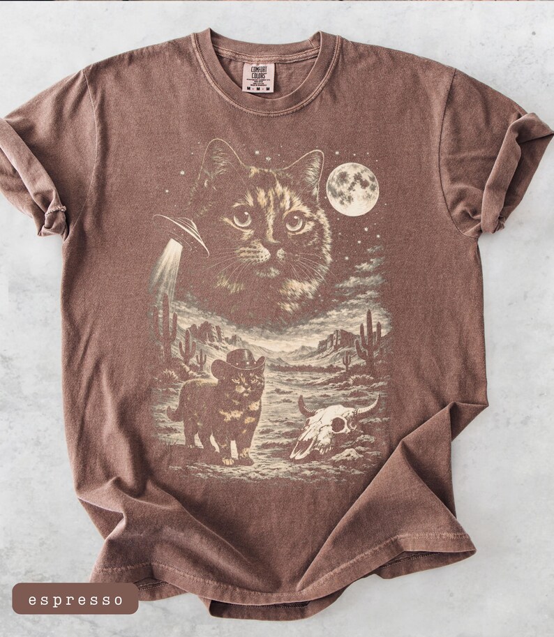 Pu&ograve; includere: T-shirt color espresso con grafica a tema gatto. Il design include un grande volto di gatto, un UFO, un gatto cowboy, un paesaggio desertico e una luna piena. La maglietta ha girocollo e maniche corte.