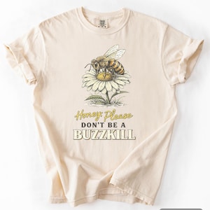 May include: Ivory-coloured t-shirt with a detailed illustration of a bee on a daisy. The text "Honey Please Don't Be A Buzzkill" is printed below the graphic. The shirt has a classic crew neck and short sleeves.