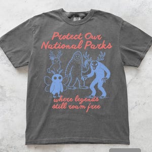 May include: A dark gray t-shirt with the text "Protect Our National Parks" in red. Below the text are blue illustrations of mythical creatures. The phrase "where legends still roam free" is printed in red below the illustrations.