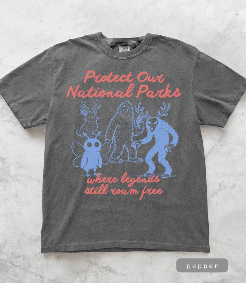 May include: A dark gray t-shirt with the text "Protect Our National Parks" in red. Below the text are blue illustrations of mythical creatures. The phrase "where legends still roam free" is printed in red below the illustrations.