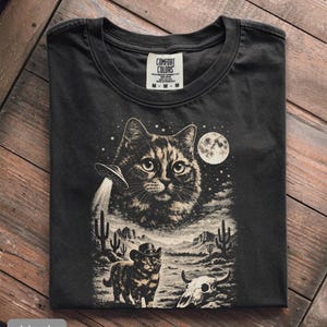Pu&ograve; includere: T-shirt nero con grafica a tema gatto. Il design include un grande volto di gatto, un UFO, un gatto con un cappello da cowboy, un paesaggio desertico e una luna piena. La maglietta &egrave; etichettata "Comfort Colors".