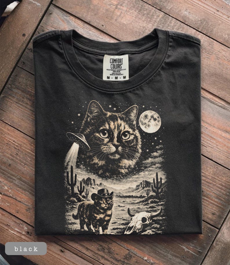 Pu&ograve; includere: T-shirt nero con grafica a tema gatto. Il design include un grande volto di gatto, un UFO, un gatto con un cappello da cowboy, un paesaggio desertico e una luna piena. La maglietta &egrave; etichettata "Comfort Colors".