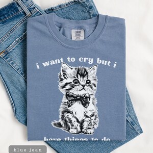 May include: A blue t-shirt with the text "i want to cry but i have things to do" and a black and white illustration of a kitten wearing a bow tie. The shirt is folded on top of a pair of blue jeans.