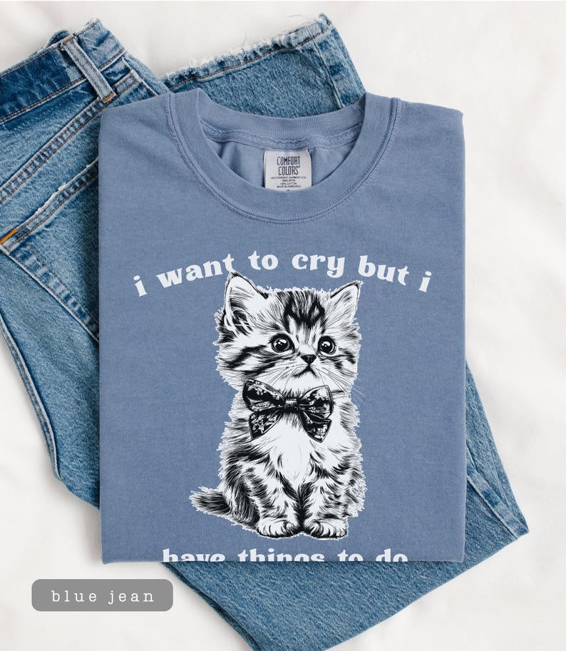 May include: A blue t-shirt with the text "i want to cry but i have things to do" and a black and white illustration of a kitten wearing a bow tie. The shirt is folded on top of a pair of blue jeans.