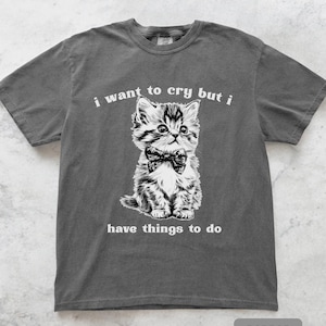 May include: Pepper-colored t-shirt featuring a black and white illustration of a kitten wearing a bow tie. The text "i want to cry but i have things to do" is printed above and below the kitten.