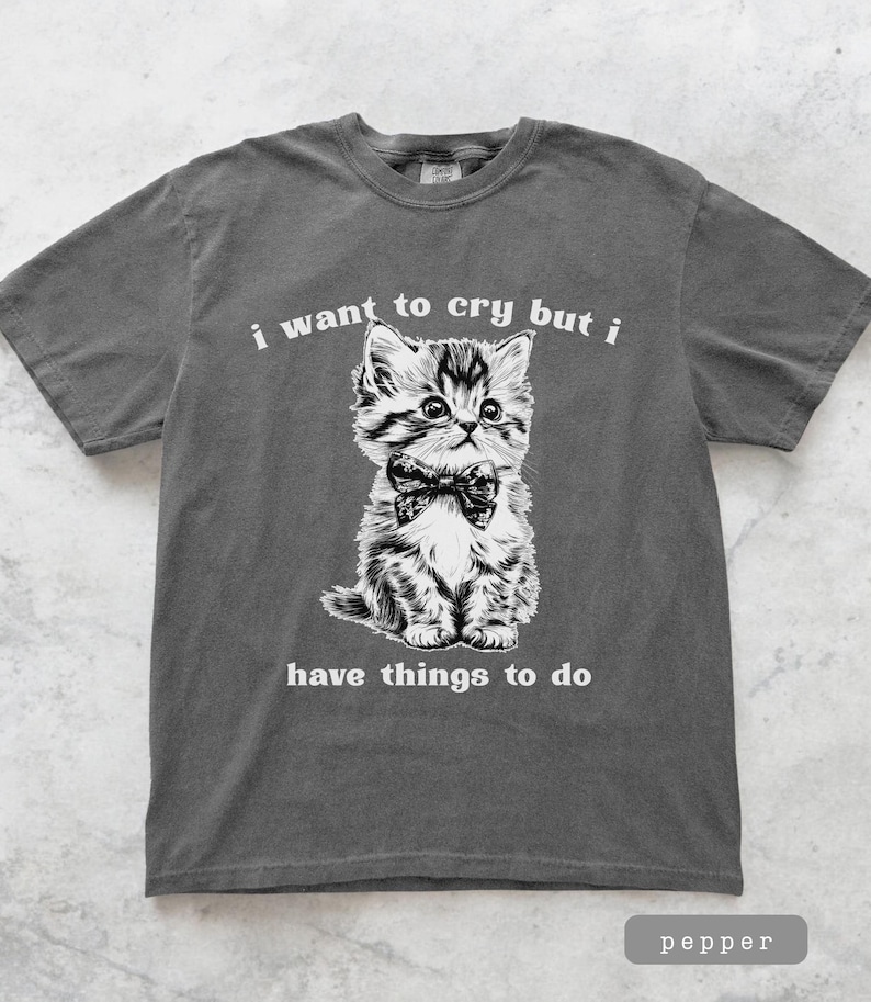 May include: Pepper-colored t-shirt featuring a black and white illustration of a kitten wearing a bow tie. The text "i want to cry but i have things to do" is printed above and below the kitten.