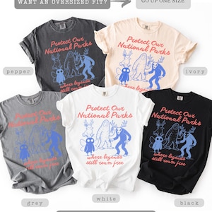 May include: A collection of soft-washed t-shirts in various colors, including pepper, ivory, grey, white, and black. Each shirt features the text "Protect Our National Parks" and a graphic of whimsical creatures. The shirts are labeled as "soft-washed & comfy."