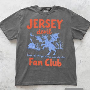 May include: Pepper-colored t-shirt with a graphic featuring the text "JERSEY devil Fan Club." The design includes a blue illustration of a mythical creature with wings, a house, and the words "lover of strange stories and wild place."