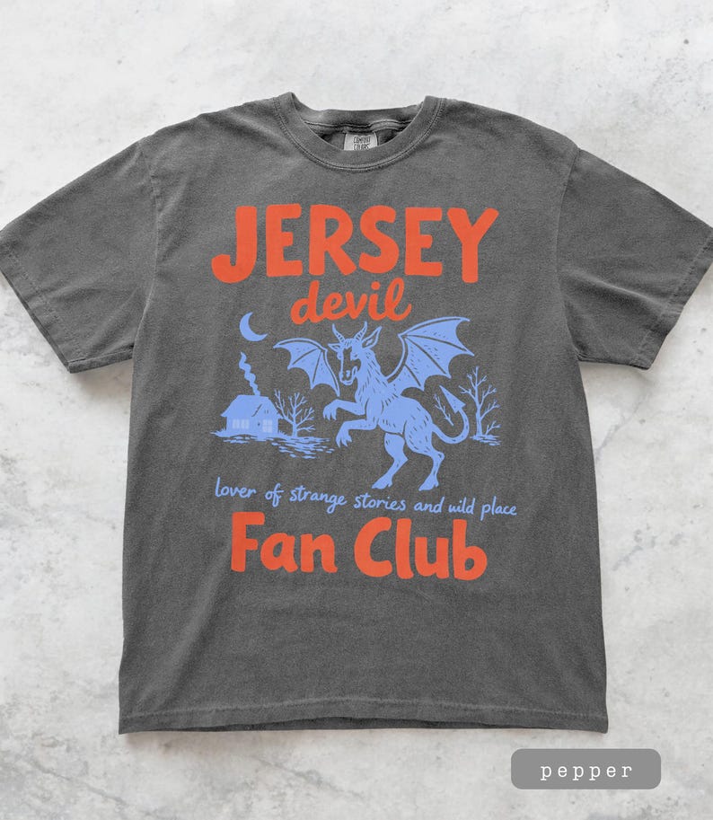 May include: Pepper-colored t-shirt with a graphic featuring the text "JERSEY devil Fan Club." The design includes a blue illustration of a mythical creature with wings, a house, and the words "lover of strange stories and wild place."