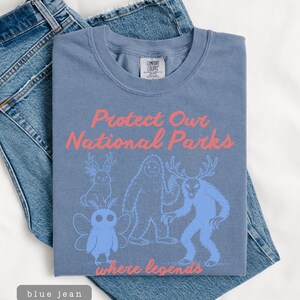 May include: A blue t-shirt with the text "Protect Our National Parks" in pink, along with illustrations of mythical creatures. The shirt is laid on top of blue jeans.