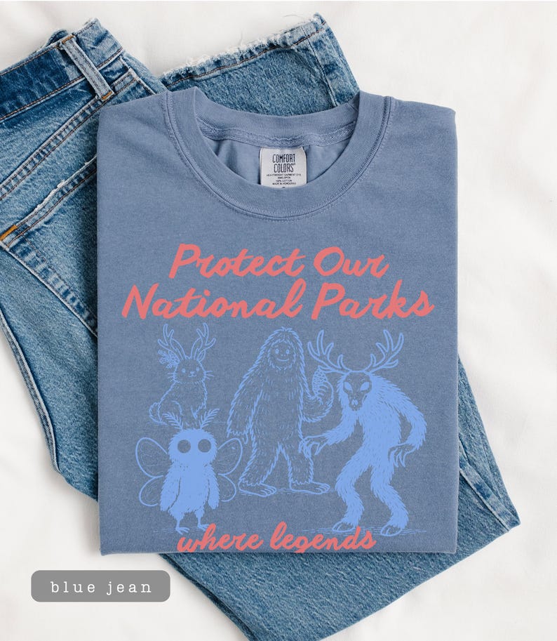 May include: A blue t-shirt with the text "Protect Our National Parks" in pink, along with illustrations of mythical creatures. The shirt is laid on top of blue jeans.