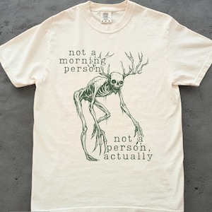 Può includere: T-shirt color avorio con un'illustrazione verde di una creatura scheletrica con corna. Il testo "not a morning person" e "not a person, actually" è stampato sopra e sotto la creatura. La maglietta è in materiale morbido.