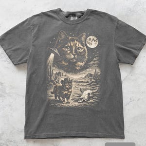 Pu&ograve; includere: T-shirt grigio scuro con grafica a tema gatto. Il design include un grande volto di gatto, un UFO, un gatto con un cappello da cowboy, un teschio e un paesaggio desertico con cactus sotto la luna piena. La parola "pepper" &egrave; in basso.