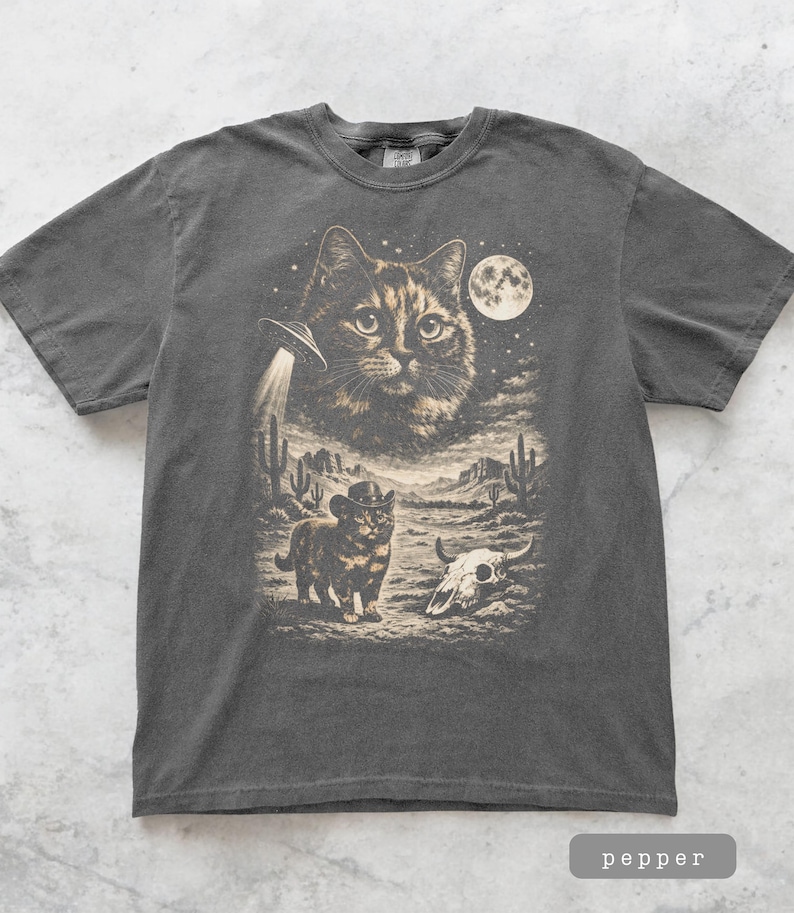 Pu&ograve; includere: T-shirt grigio scuro con grafica a tema gatto. Il design include un grande volto di gatto, un UFO, un gatto con un cappello da cowboy, un teschio e un paesaggio desertico con cactus sotto la luna piena. La parola "pepper" &egrave; in basso.