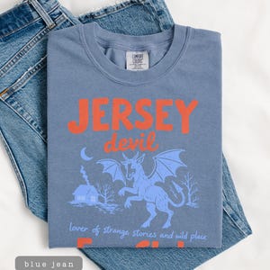 May include: A blue t-shirt with the words "JERSEY devil" in orange, featuring a blue illustration of a horned creature with wings. The shirt is folded on top of blue jeans.