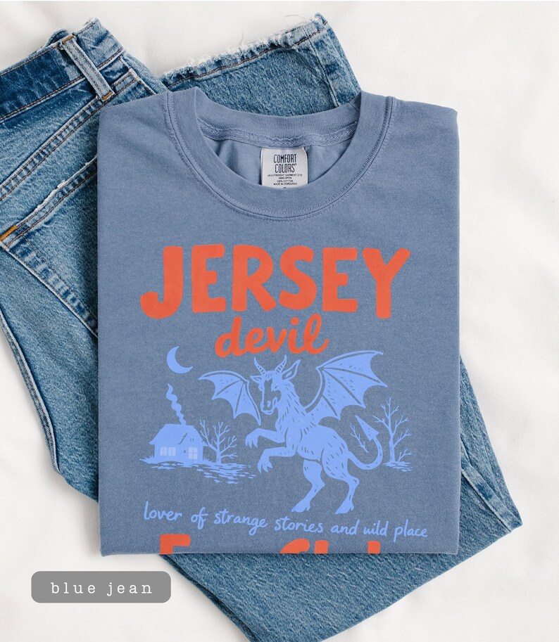 May include: A blue t-shirt with the words "JERSEY devil" in orange, featuring a blue illustration of a horned creature with wings. The shirt is folded on top of blue jeans.