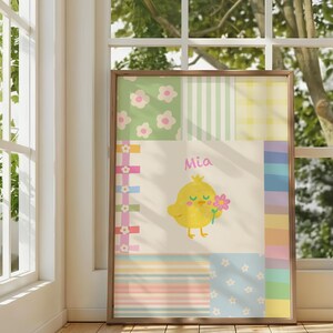 Könnte beinhalten: Ein gerahmter Wandkunstdruck mit einem cremefarbenen Mittelfeld, das ein gelbes Küken mit einer rosa Blume und den in Rosa geschriebenen Namen Mia zeigt. Das Design ist von einem Patchwork-Rand mit pastellfarbenen Blumen-, Streifen- und Karomustern umgeben.