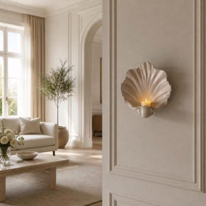 May include: A wall-mounted candle holder shaped like a large, off-white seashell with a scalloped edge. The holder features a central indentation designed to securely hold a single lit pillar candle, casting a warm glow against the textured surface.