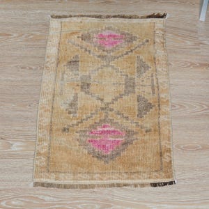Puede incluir: Una alfombra rectangular beige con un patr&oacute;n geom&eacute;trico. El dise&ntilde;o presenta formas de diamante en rosa y gris, con un diamante central y formas de diamante en cada extremo. La alfombra tiene un borde con flecos y est&aacute; colocada sobre un suelo de madera clara.