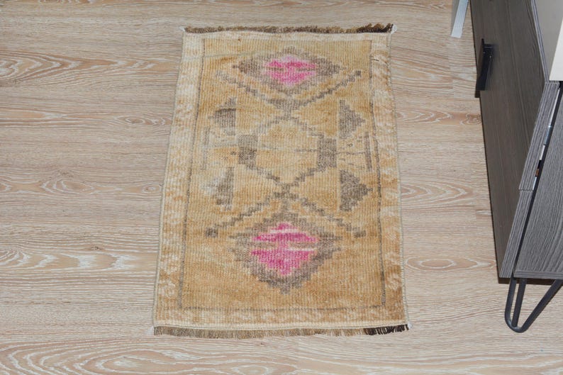 Puede incluir: Una alfombra rectangular beige con un patr&oacute;n geom&eacute;trico. El dise&ntilde;o presenta formas de diamante en rosa y gris, con un diamante central y formas de diamante en cada extremo. La alfombra tiene un borde con flecos y est&aacute; colocada sobre un suelo de madera clara.