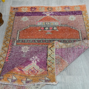 Puede incluir: Una alfombra rectangular de estilo vintage con una esquina doblada. La alfombra presenta un diseño central en tonos naranja, morado y rosa, con motivos florales y geométricos. El borde es una mezcla de naranja y amarillo.