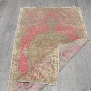 Könnte beinhalten: Ein rechteckiger Teppich im Vintage-Stil mit verblasstem Aussehen. Der Teppich hat ein zentrales Design in Rot- und Olivgrüntönen auf beigem Hintergrund. Die Kanten sind leicht ausgefranst, was seinen antiken Charme unterstreicht.